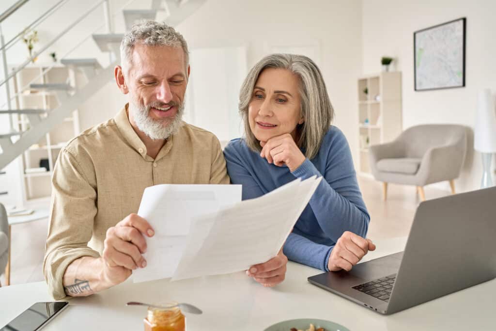 middle aged couple reading retirement papers and 401k limits using laptop at home.