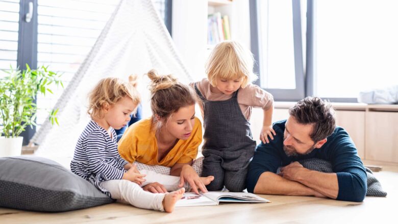 young family with two small children indoors in bedroom reading a book.