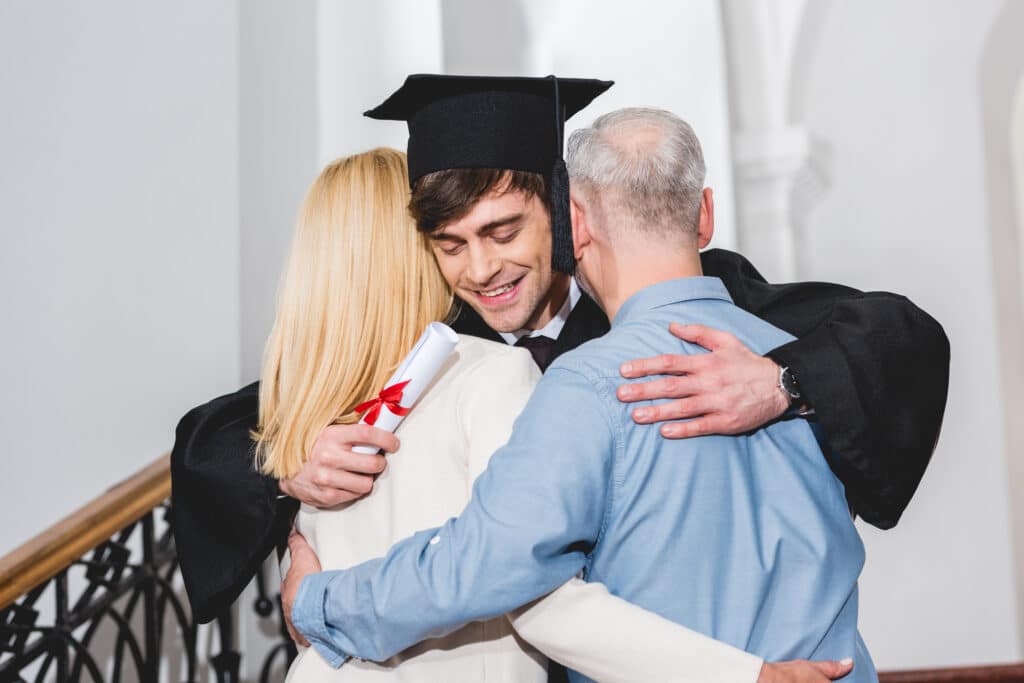 cheerful son in graduation cap holding diploma while hugging parents