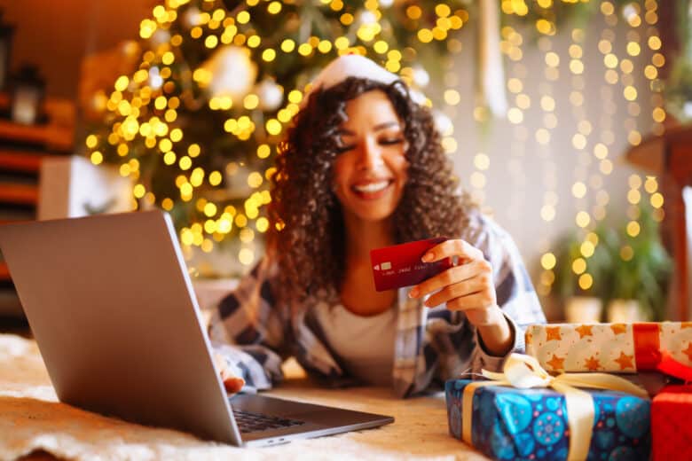 smiling african american woman using laptop, sitting near christmas tree with card, shopping online